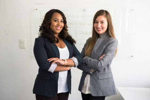 Two diverse women in dress clothes wmiling with arms crossed in front of white board. Photo by Christina @ wocintechchat on Unsplash